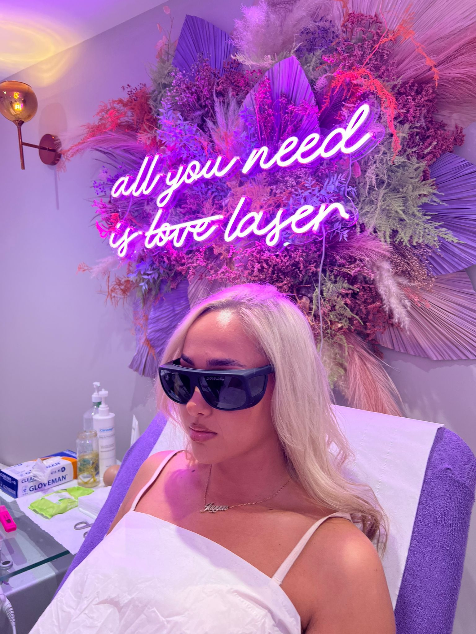 Woman receiving Laser Genesis treatment at Escape Aesthetics wearing protective glasses with "All you need is love laser" neon sign in the background.